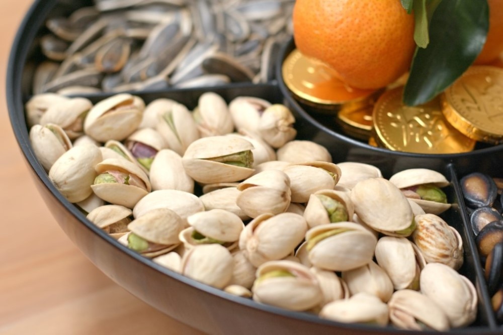  A traditional Lunar New Year snack tray, featuring pistachios, sunflower seeds, an orange with leaves, and gold chocolate coins, symbolizing prosperity and luck